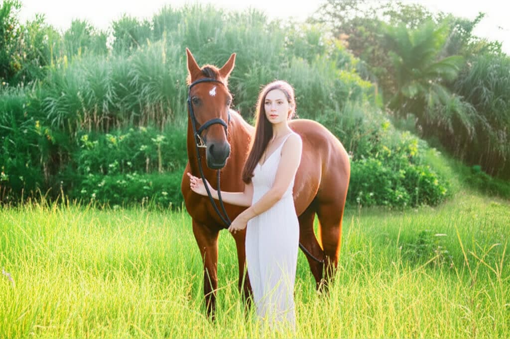 A woman standing calmly beside a horse in long grass